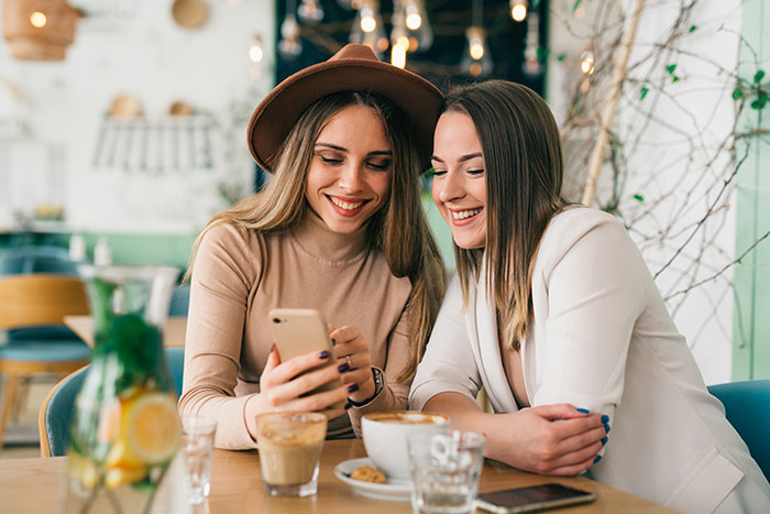 Two women sitting at a café table, smiling and looking at a smartphone.
