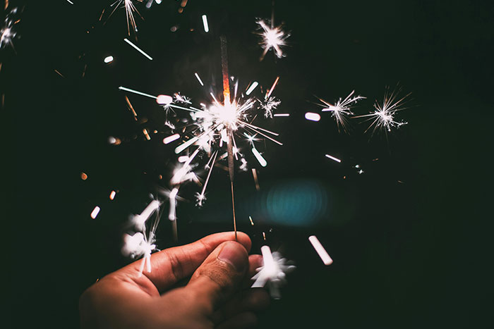 A hand holding a lit sparkler with bright sparks on a dark background.