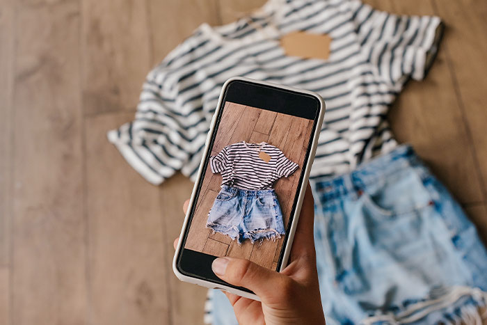 Hand holding a smartphone displaying a striped t-shirt and denim shorts, with the same outfit on the wooden floor in the background.