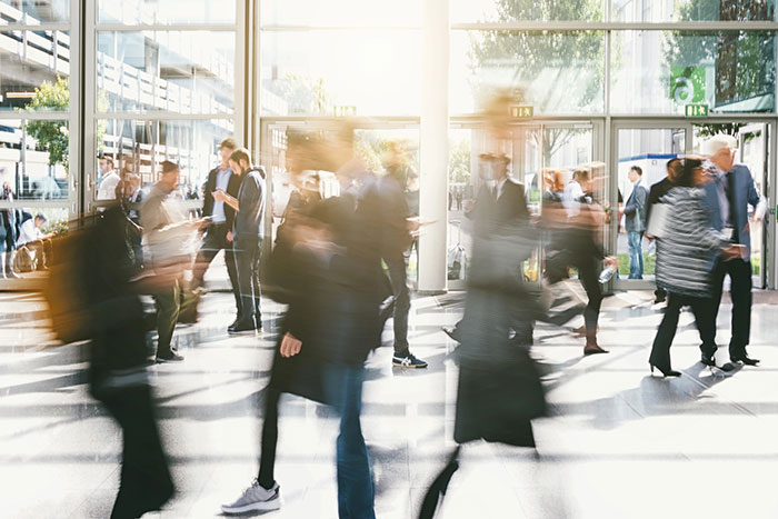 A busy indoor walkway with blurred people in motion and large windows letting in sunlight.