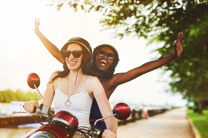 Two people enjoying a scooter ride on a sunny day, both wearing helmets and sunglasses, with trees lining the road.