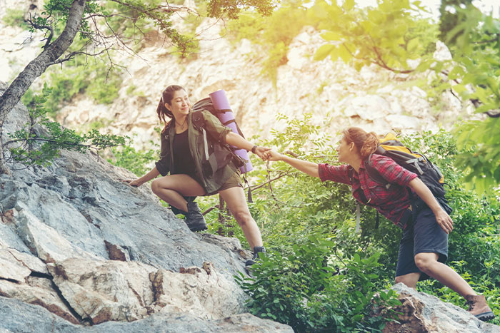 Two women hiking on rocky terrain, one helping the other up a steep section.