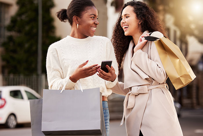 Two women smiling and carrying shopping bags in an outdoor urban setting.